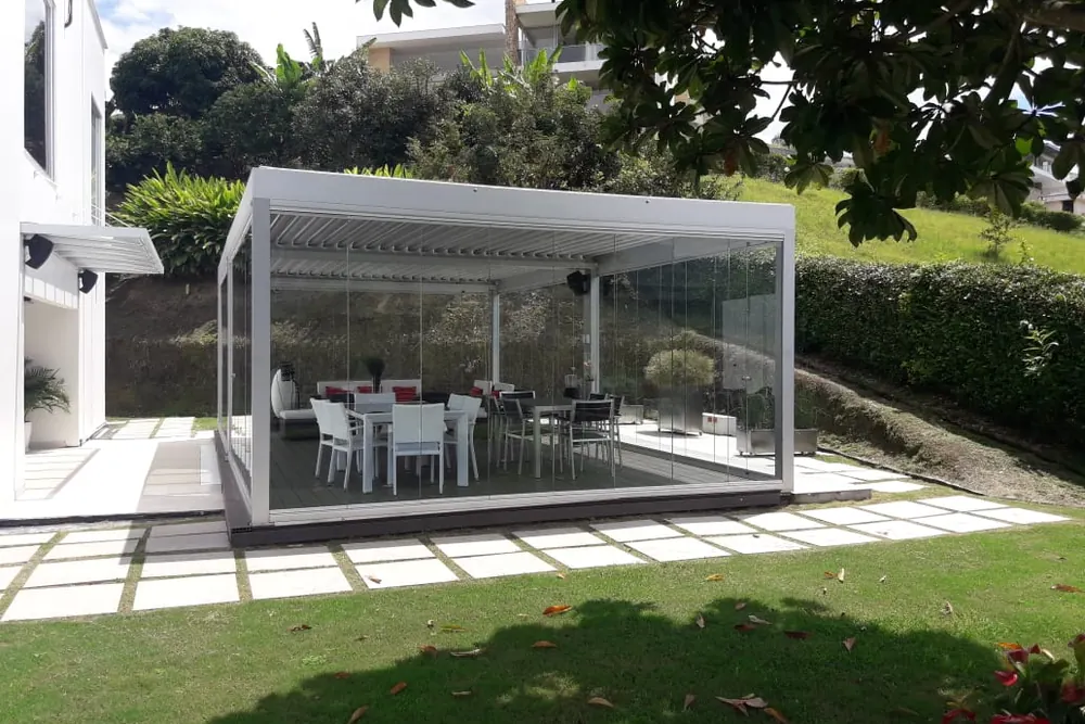 Comedor exterior cerrado con paneles de vidrio Slidinglass bajo una pérgola de aluminio blanco