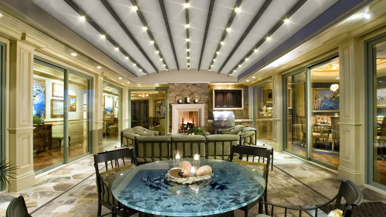 Pérgola retráctil PergoDom entre muros sobre un patio comedor, con la lona curva cerrada e iluminación integrada de noche