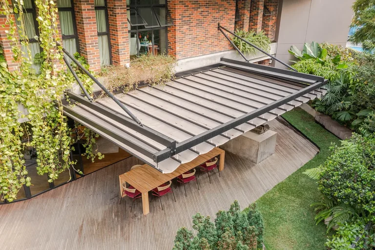 Pérgola retráctil PergoFlex semiabierta sobre el deck en madera del patio interior del Edificio Gulungo, con mesa de comedor larga y jardín