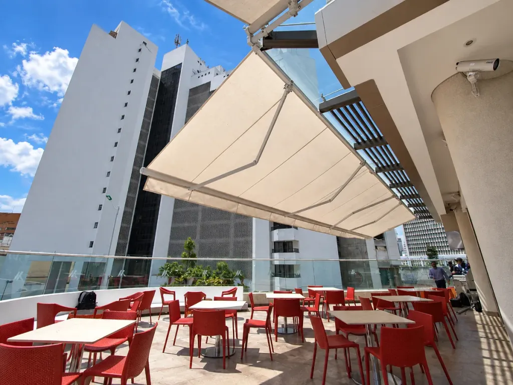 Toldo Supersombralinas con los brazos a la vista sobre la terraza de un edificio, con mesas y sillas rojas