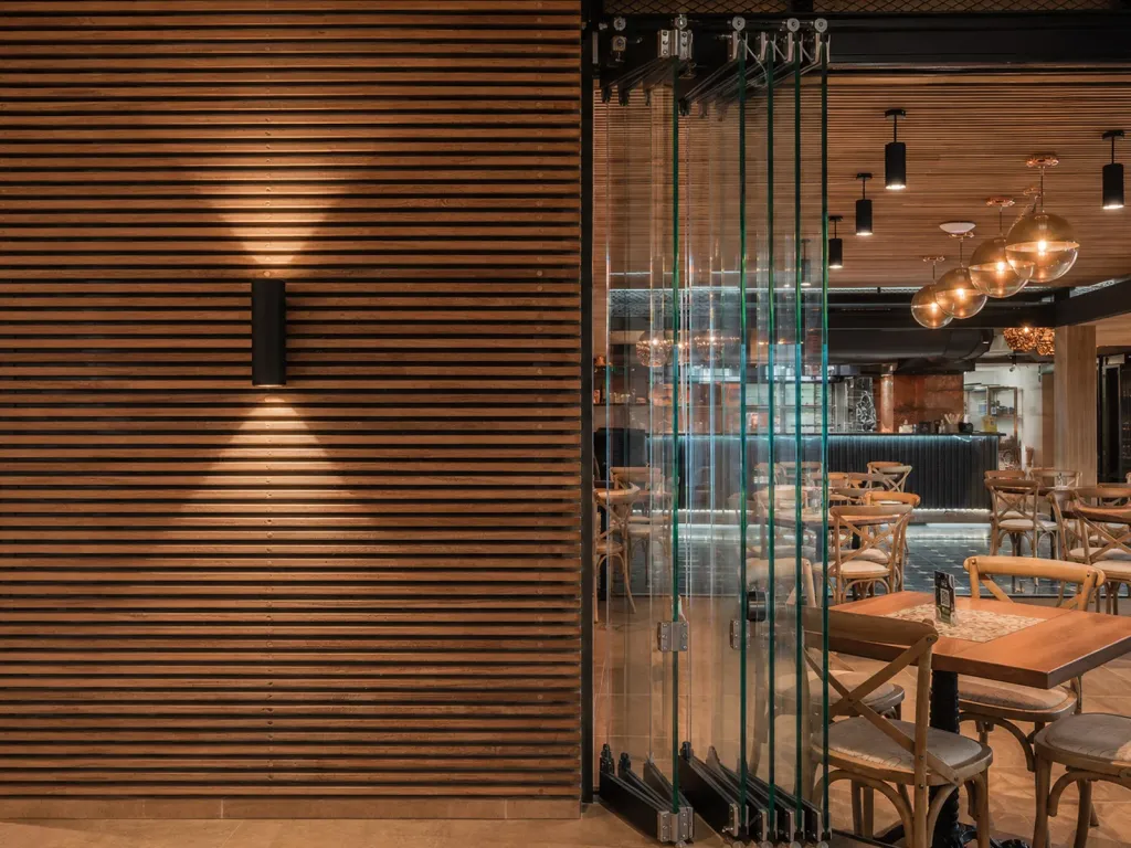 Cerramiento FlexiGlass de vidrio plegado junto a un muro de listones de madera en un restaurante