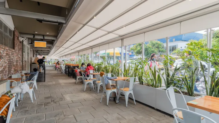 Pérgola retráctil cerrada sobre la terraza de mesas con jardineras de Carulla Oviedo en Medellín