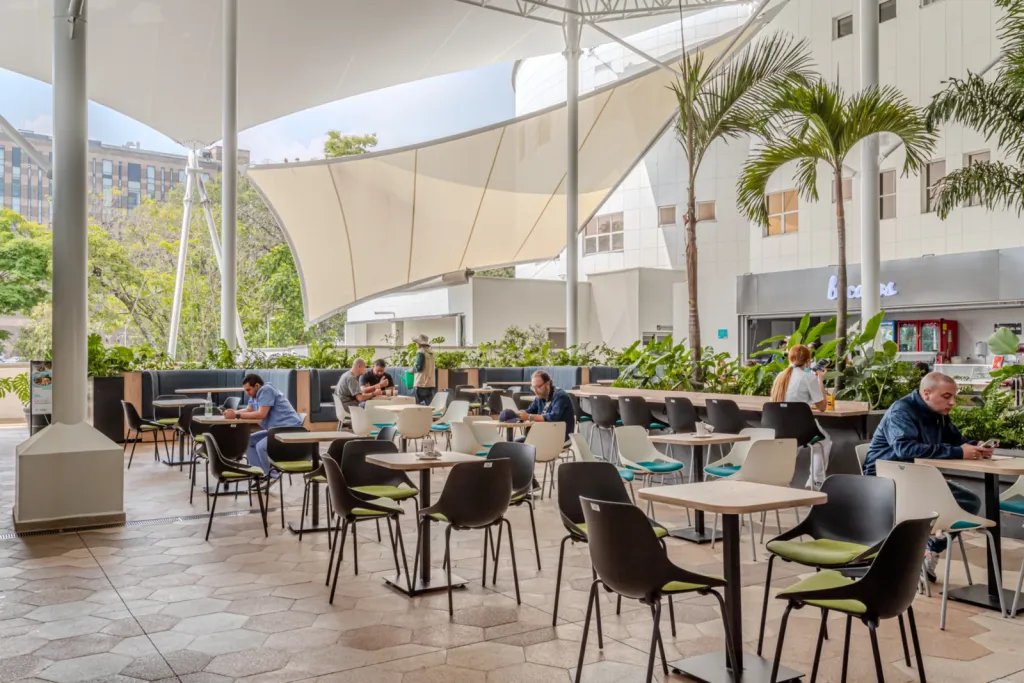 Zona de cafetería bajo la membrana tensada de la Clínica Las Américas con mesas y palmas
