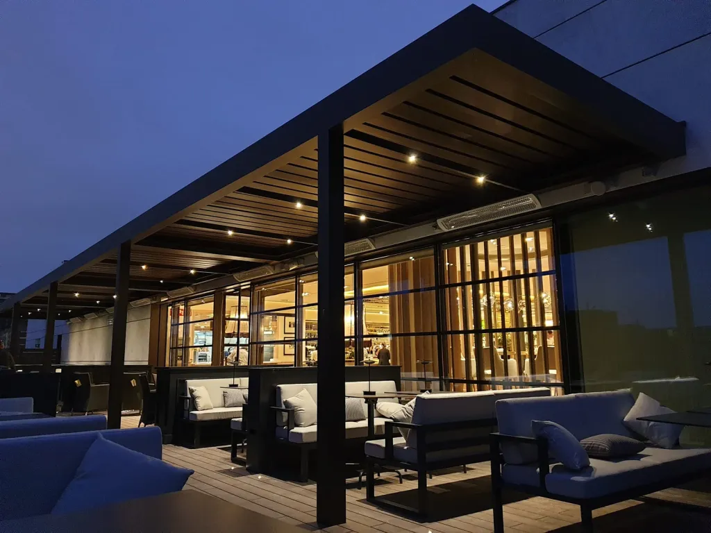 Pérgola bioclimática PergoLam sobre la terraza de un restaurante con fachada de vidrio, al anochecer