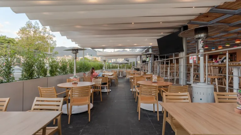 Terraza larga de Archies 116 bajo una pérgola retráctil de tela, con mesas de madera y calentadores