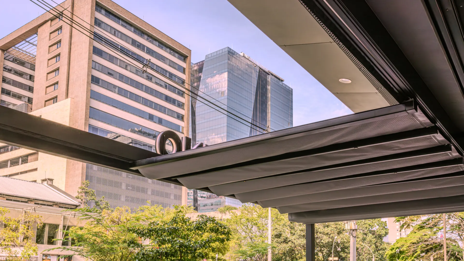 Pérgola retráctil de lona recogida sobre la terraza de Pergamino Café en Laureles, Medellín, con edificios de oficinas al fondo