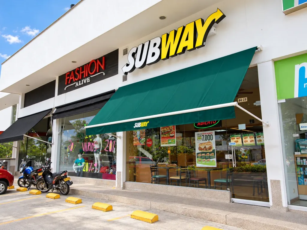Toldo verde de brazos proyectantes extendido sobre la fachada de Subway junto a los locales vecinos