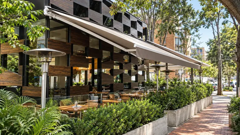 Toldo retráctil extendido sobre la terraza de Creppes & Waffles, entre jardineras y la acera de ladrillo